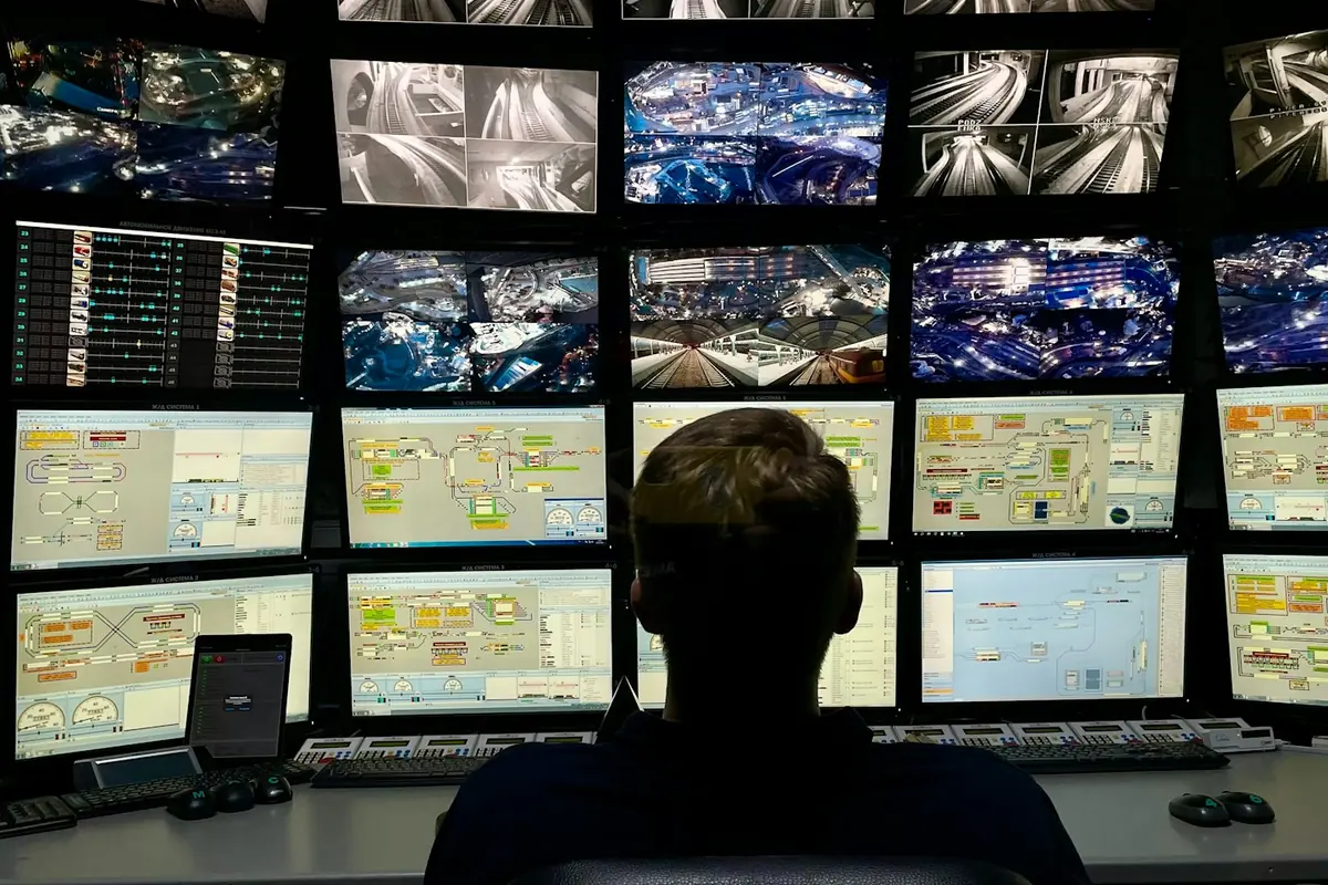 Sala de monitoreo a oscuras con un muro de pantallas mostrando cámaras de vía y tableros de operación.